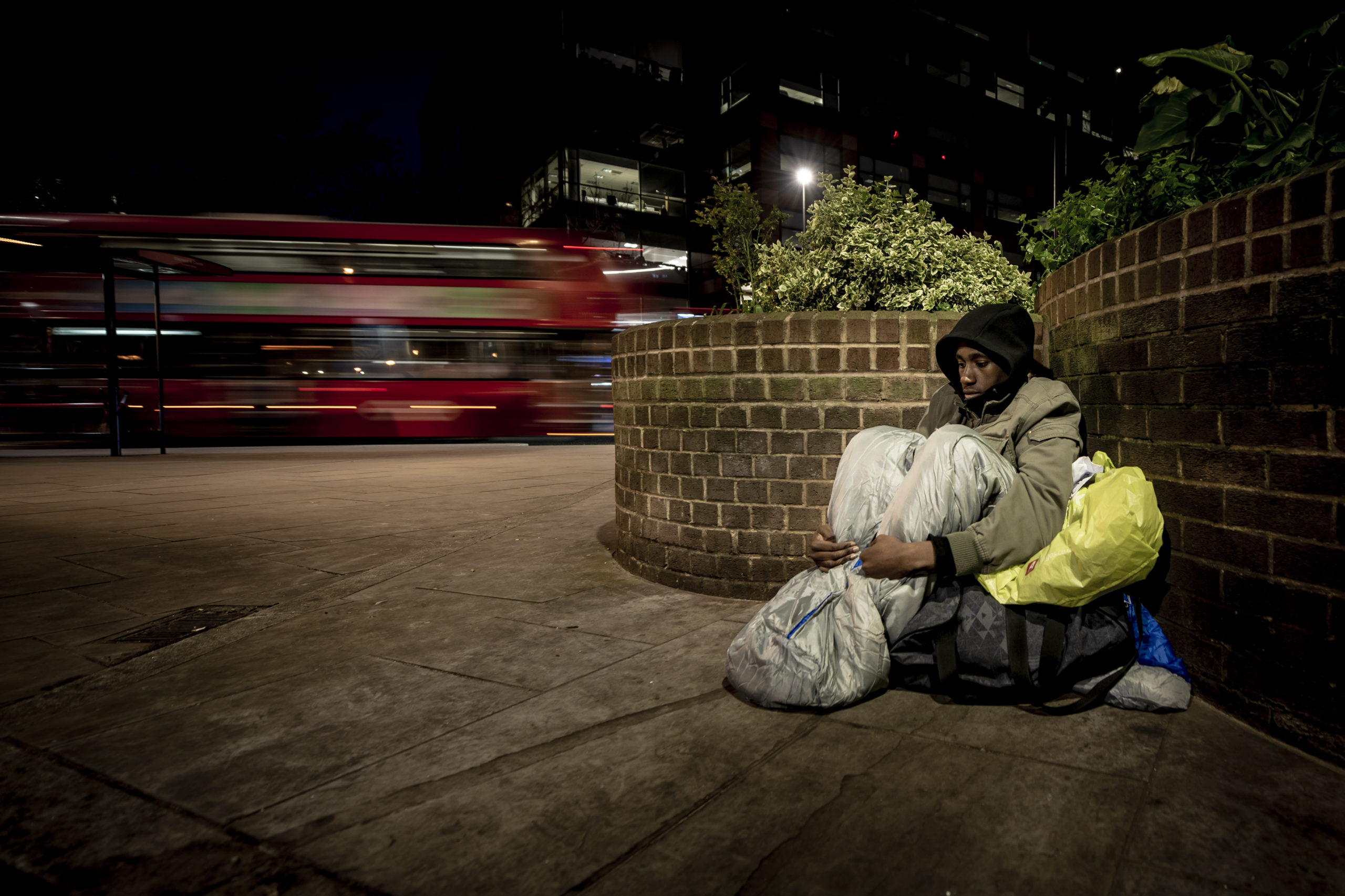 Homeless man sitting on the ground, resting against a brick fence. He has a few belongings next to him. Night time, outdoors and a bus driving in the background.