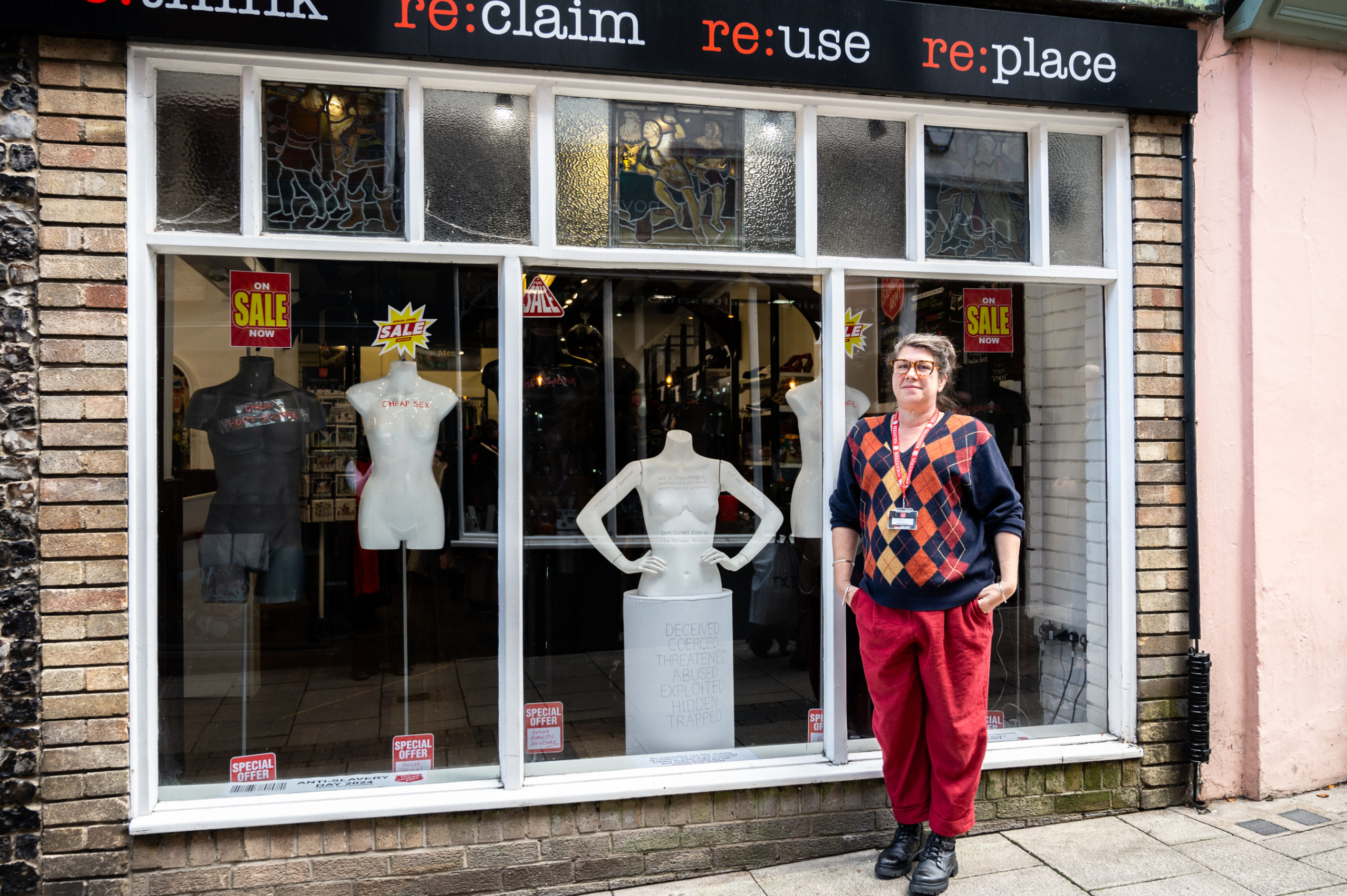 lady standing in front of window display, dressed in red trousers and chequered jumper with sleeves rolled up. Lanyard around the neck and black shoes.