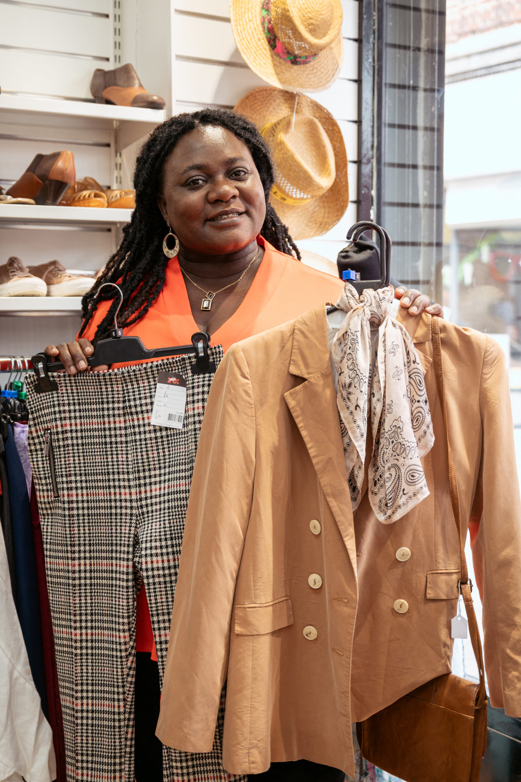 Salvation Army's Luciana holding a pair of checkered trousers in one hand, and a blazer with a scarf in the other hand. Clothing items are on hangers and there are shoes and hats in the background.