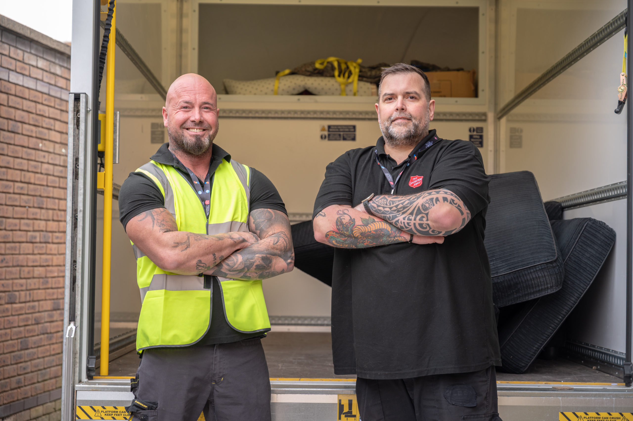 Two men with their arms folded stand at the back of a delivery van looking into the camera, both have black polo shirts, on one the Salvation Army logo is visible
