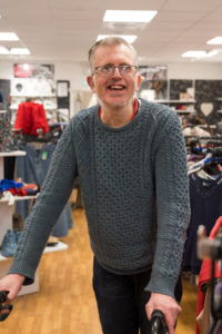 A man in a grey jumper is in the middle of the Cosham Salvation Army charity shop, smiling into the camera