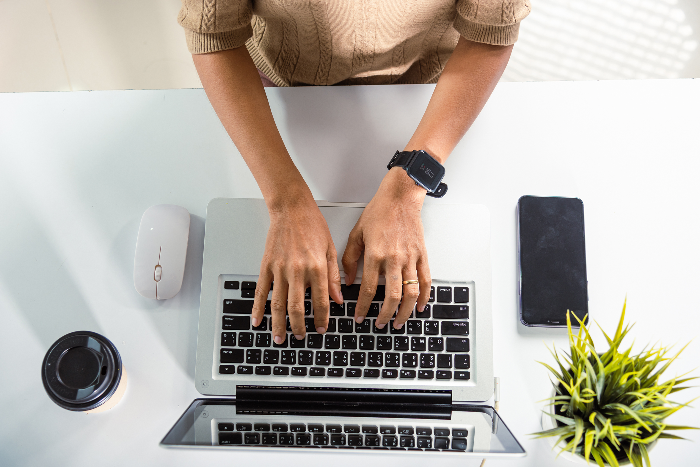 Hands typing on laptop keyboard, mouse, phone, plant, and coffee cup around the laptop.